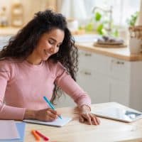 a smiling woman sits at a table and writes a letter