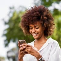 una mujer sonriente con el pelo encrespado de pie en la calle y pulsando un botón de un teléfono móvil