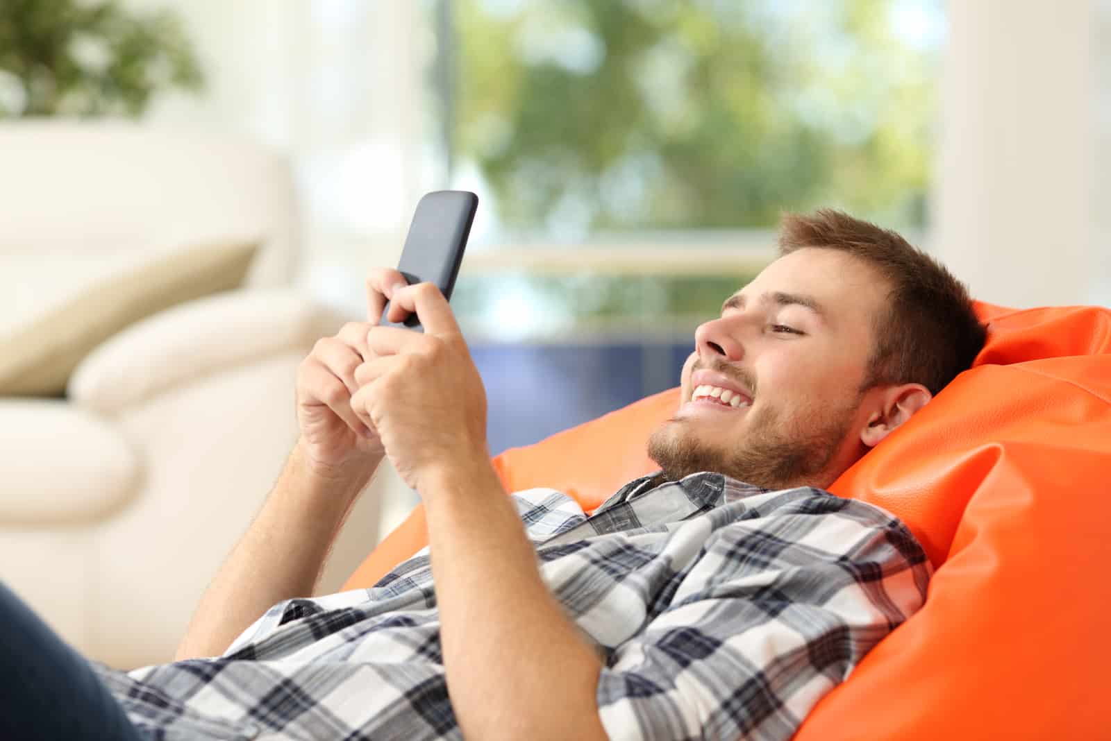 Hombre relajado usando un smartphone tumbado sobre un puf naranja