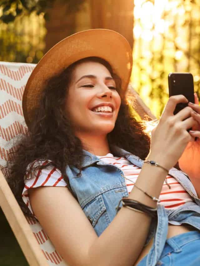 a smiling woman with a hat on her head is sitting on a chair with a phone in her hand