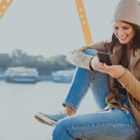 Beautiful young woman sitting by the river and using her mobile phone