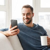 hombre sonriente con un teléfono en la mano y una taza de café sentado en el sofá