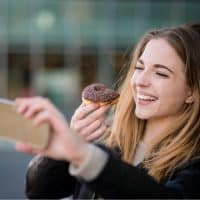 Young woman taking selfie with phone while eating chocolate