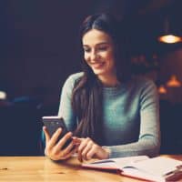 una chica sonriente con el pelo largo y negro se sienta en una mesa y utiliza un teléfono móvil