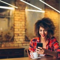 una mujer sonriente con el pelo encrespado se sienta en una mesa de una cafetería y sostiene un teléfono móvil en la mano