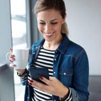 a smiling woman is standing by the window and holding a mobile phone in her hand