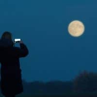 a woman stands and takes a picture of the moon with her mobile phone