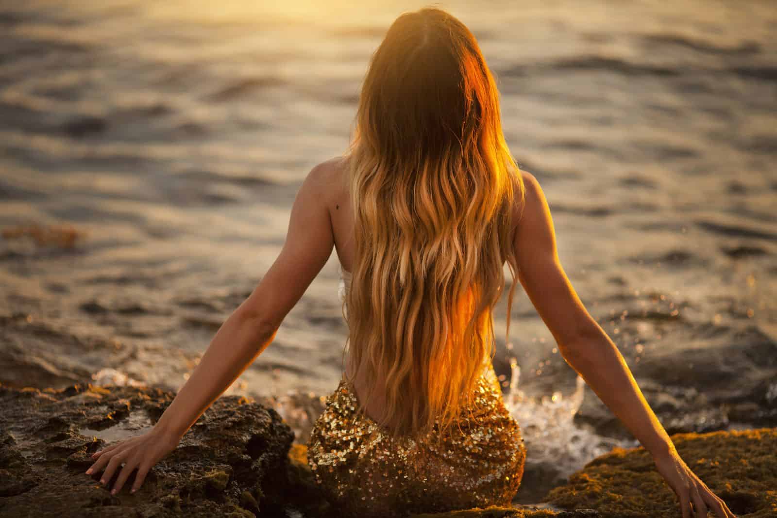 a mermaid woman sits on a rock by the water