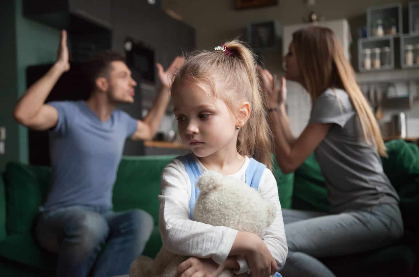 little girl holding a teddy bear while parents fight in background