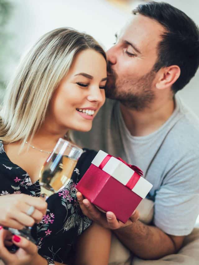 a man in grey shirt holding a red present and kissing a girl with a blonde hair