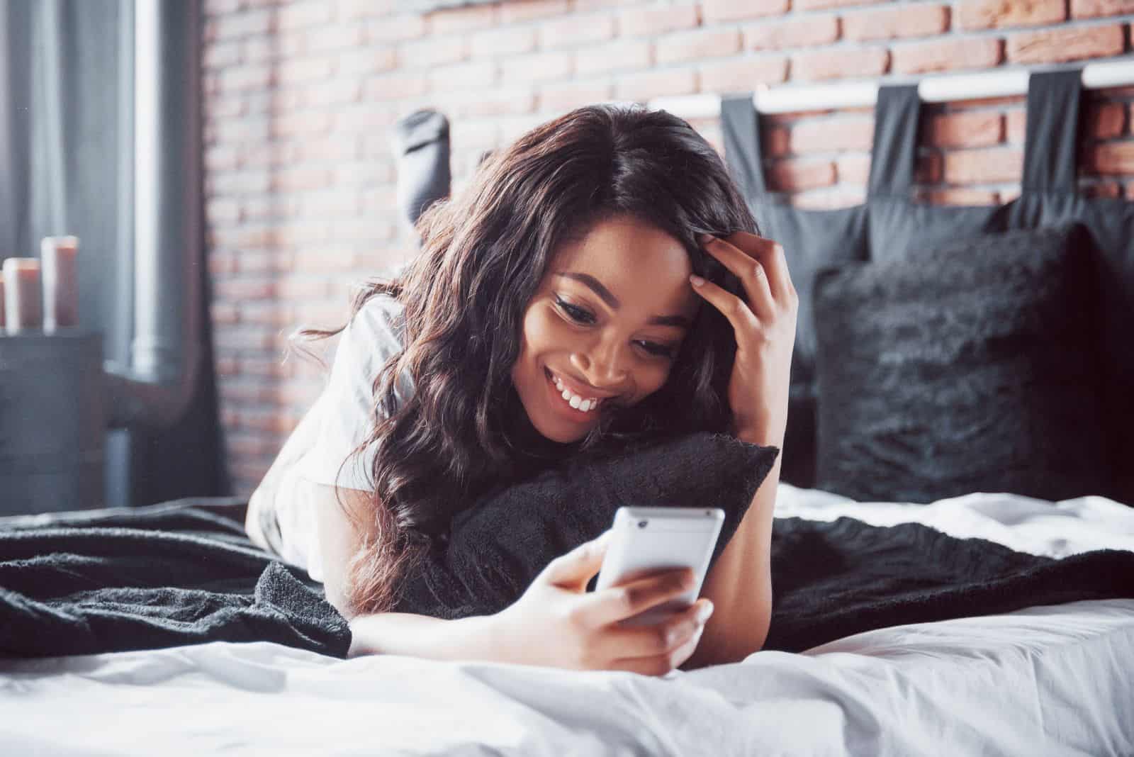 una chica sonriente está tumbada en la cama y tecleando en el teléfono