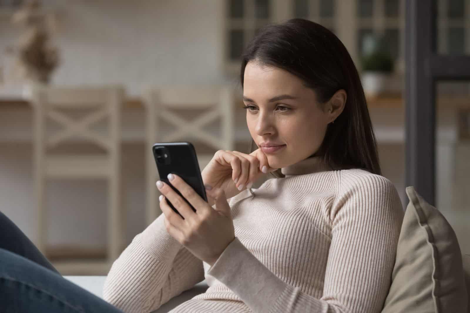 una hermosa chica está sentada en el sofá y sostiene un teléfono móvil en la mano