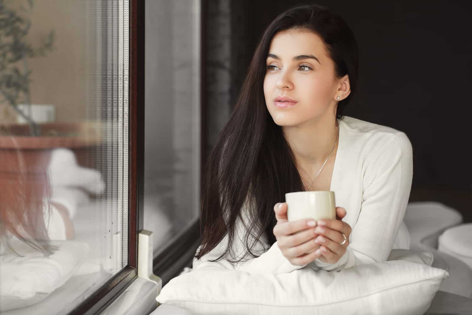 mujer con una taza de café en la mano sentada junto a una ventana