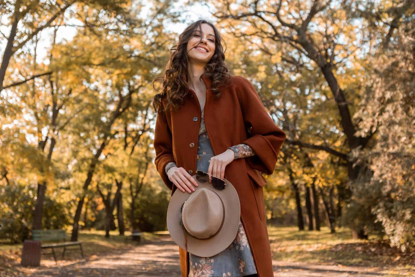 woman holding hat and sunglasses walking in park dressed in warm coat autumn