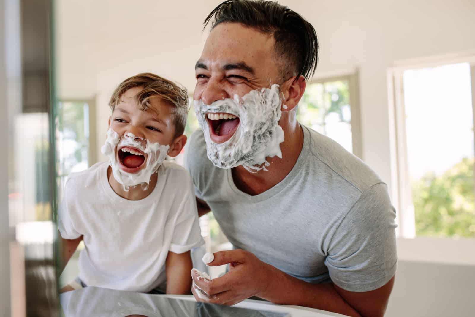 Man and little boy with shaving foam on their faces looking into the bathroom mirror and laughing