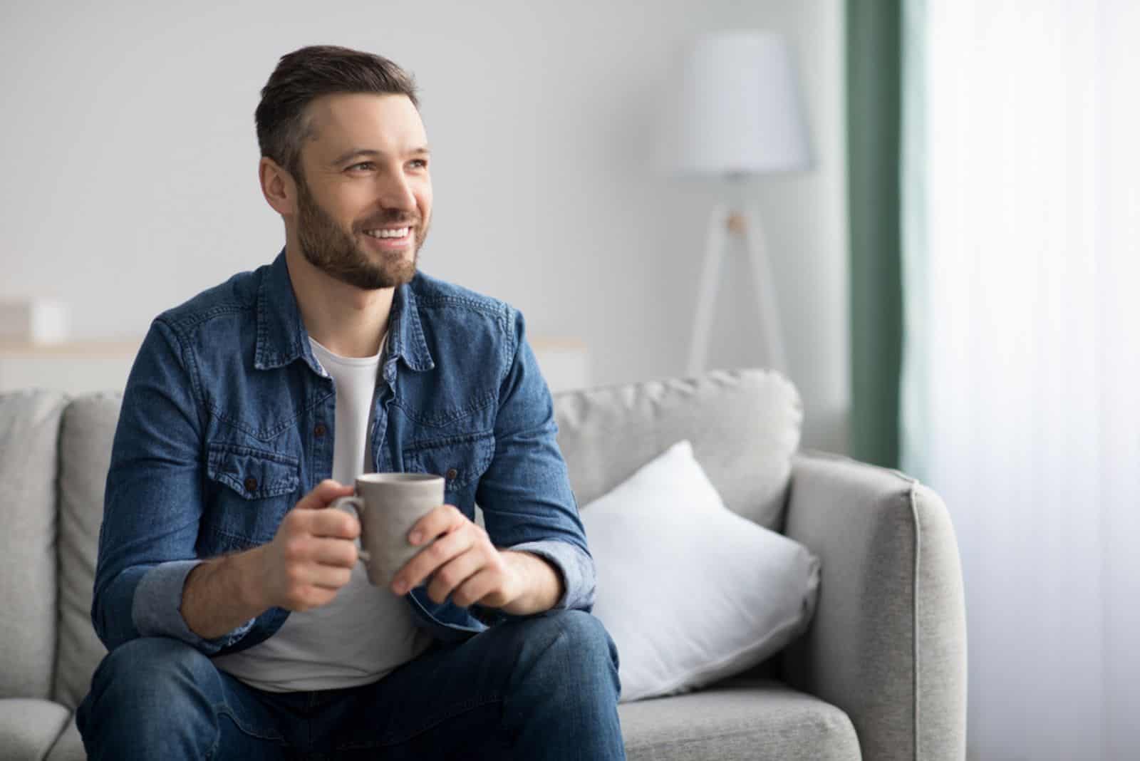 un hombre sonriente está sentado en el sofá y sostiene una taza en la mano