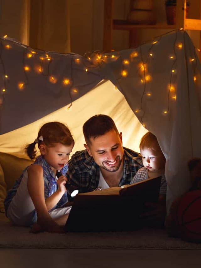 familia feliz padre e hijos leyendo un libro con una linterna en una tienda de campaña en casa