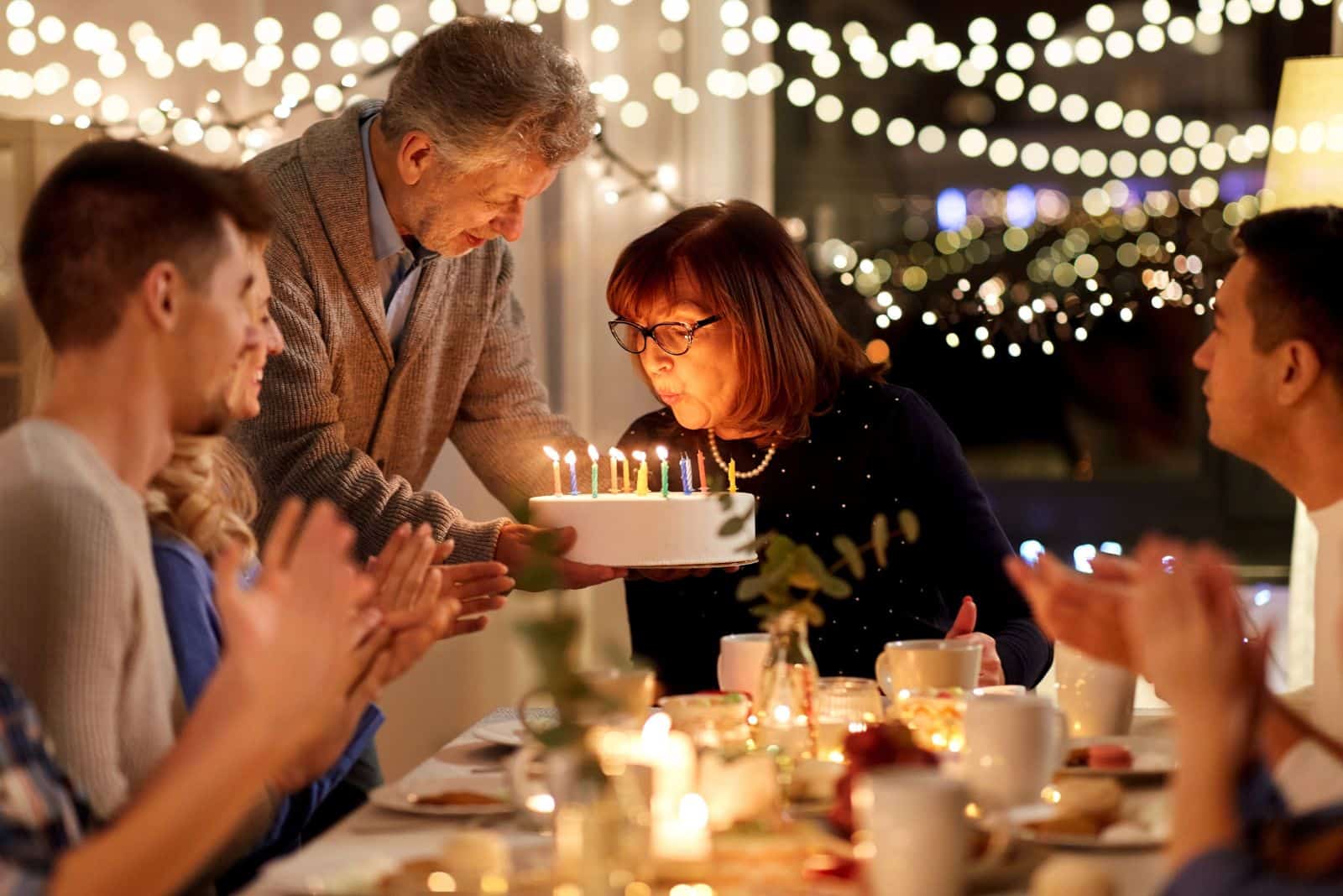 happy grandmother blowing candles on birthday cake at dinner party at home