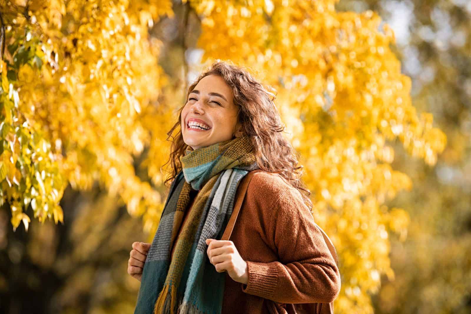 Cheerful young woman with winter scarf relaxing at park with yellow trees in background