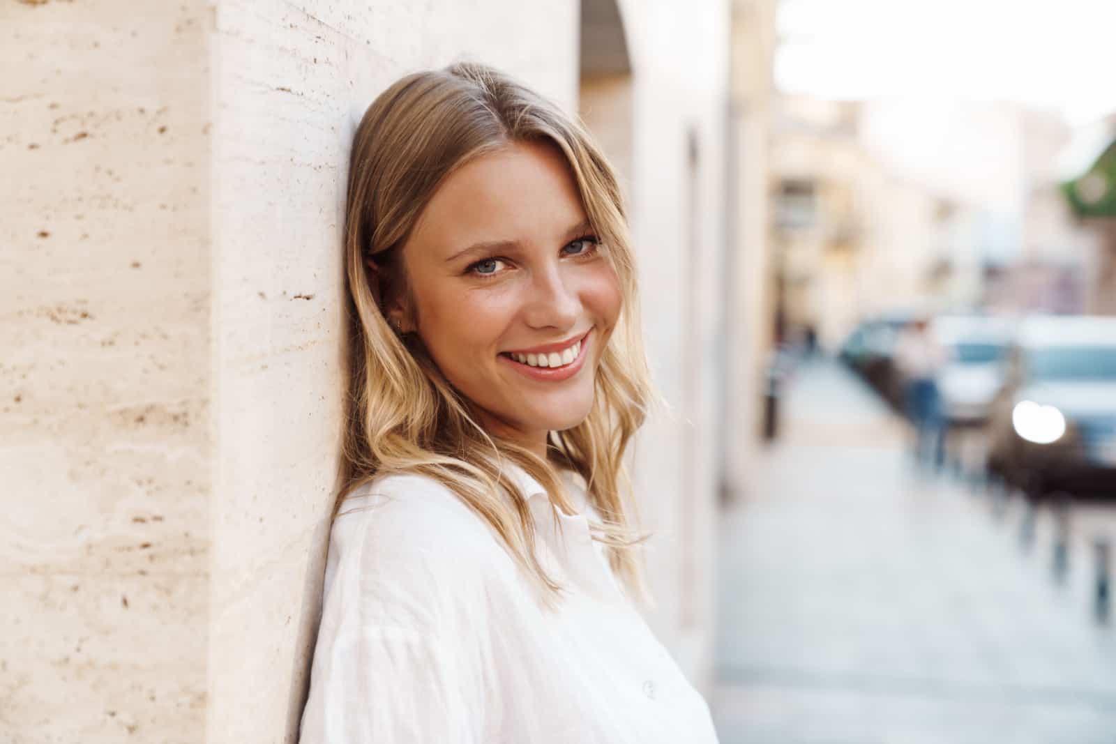 Hermosa mujer feliz sonriendo y mirando a la cámara mientras se apoya en la pared en la calle de la ciudad